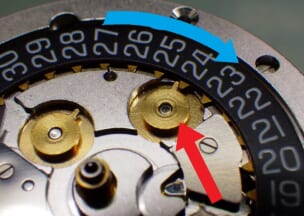 ジン 103 矢印で指した日送り車とカレンダー板が噛み合った状態です。青矢印はカレンダー板の回転方向です。